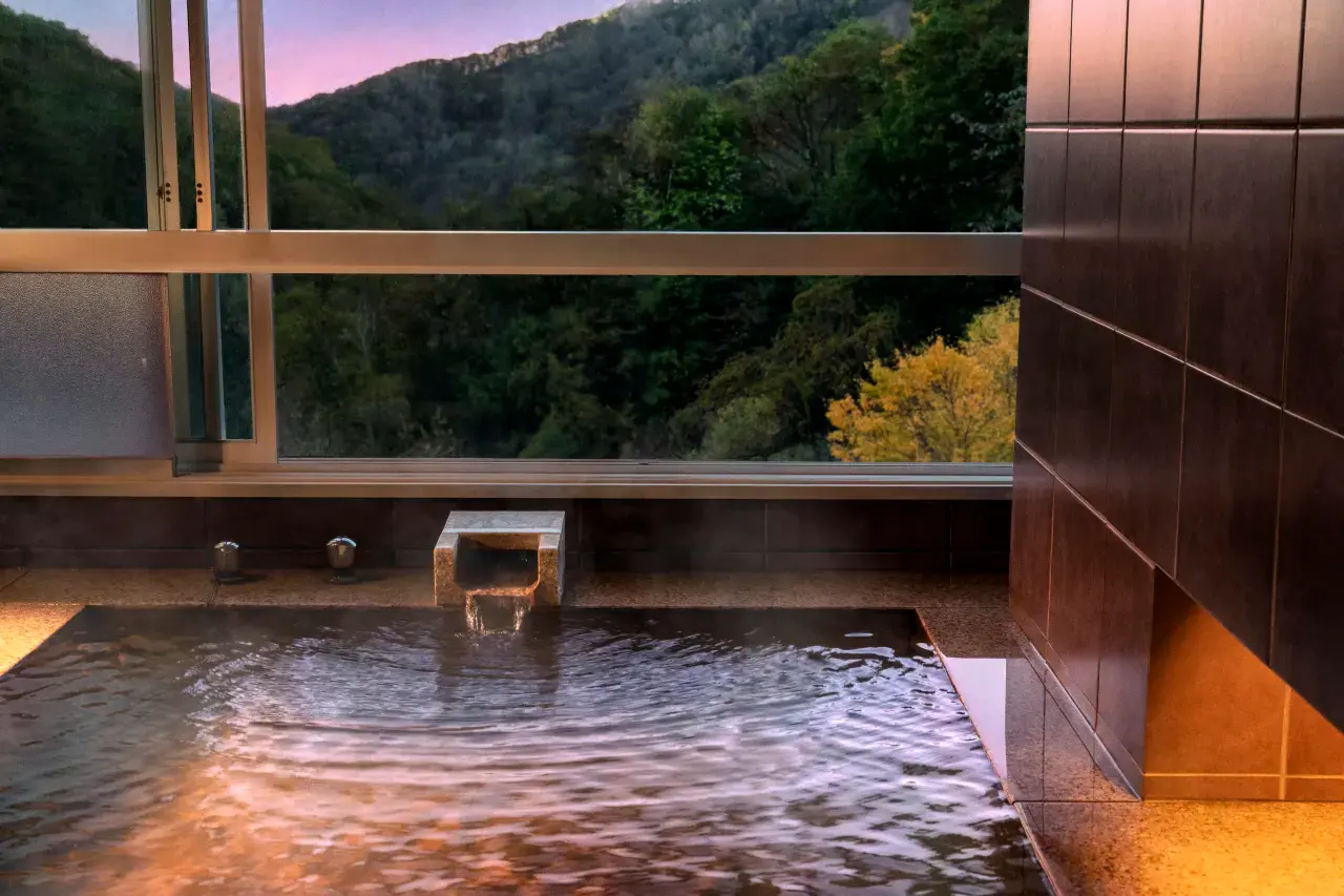 Private, warmly lit modern onsen suite at Bourou Noguchi Noboribetsu, where a sleek stone bath overlooks a dramatic view of twilight-shrouded, densely forested mountains with touches of autumn gold.